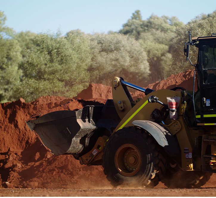 Front of a wheel loader moving red dirt on a mine site with trees in background.