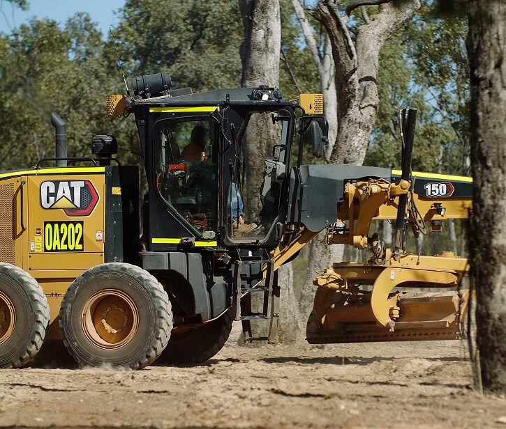 CAT motor grader working on a dirt track through a wooded area.
