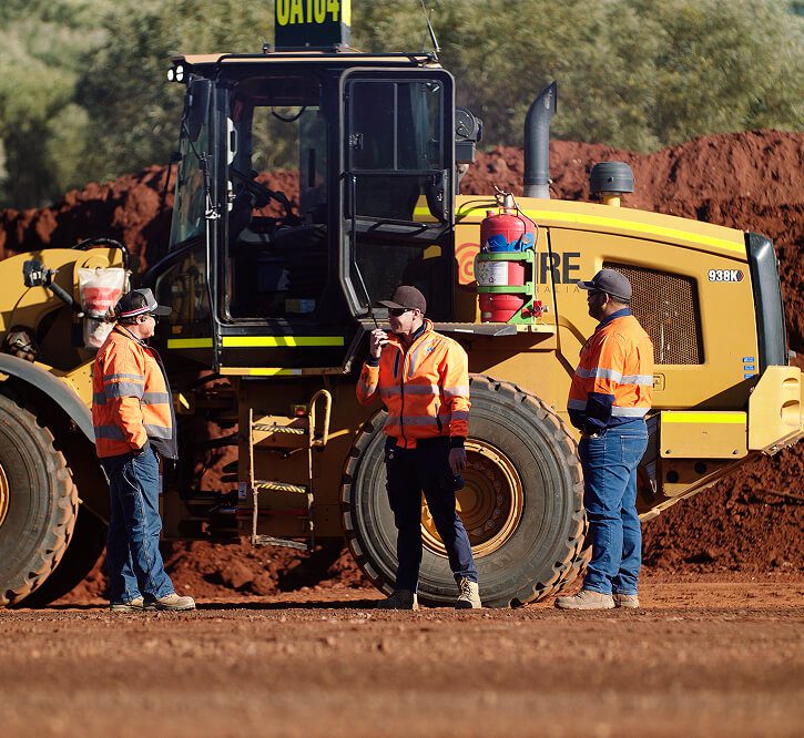 Three workers in orange hi-vis standing beside a CAT loader on a mine site.