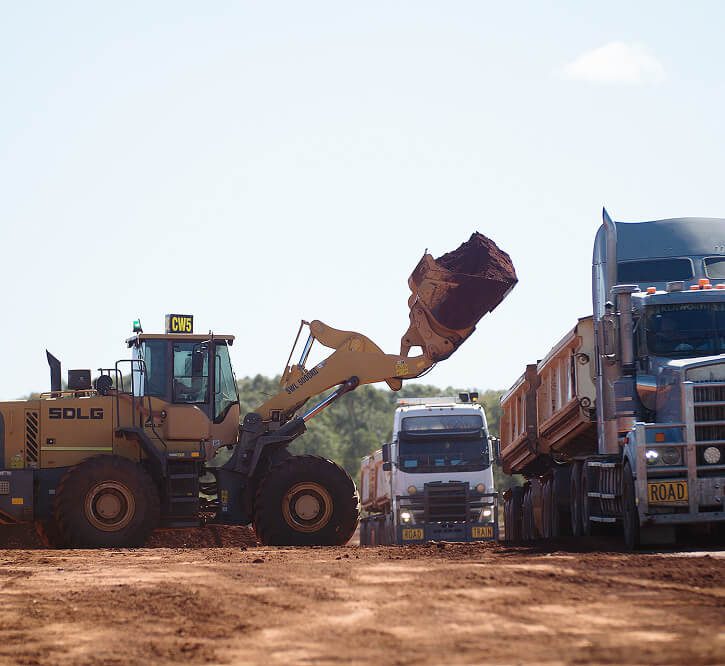 Wheel loader lifting a bucket of red dirt to load into road train trucks on a construction site.