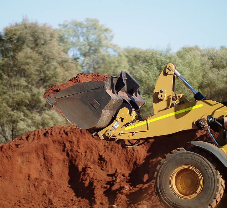 Front of a wheel loader moving red dirt on a mine site with trees in background.
