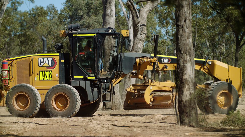 CAT motor grader working on a dirt track through a wooded area.